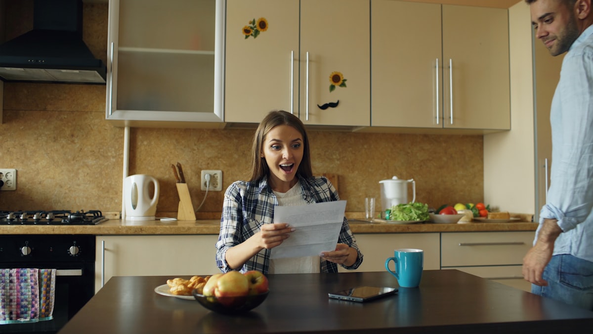 Woman reading good news from a letter at her kitchen table as her partner walks in